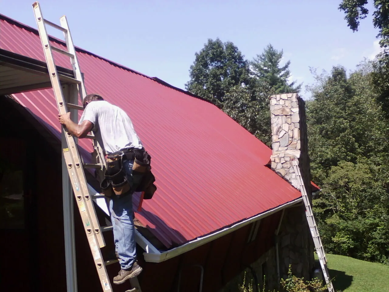 Roofer on a ladder beside a red metal roof and stone chimney — Soffit & Fascia Repair by Roofing Brothers in Barnegat